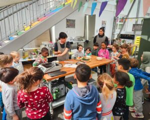 A kindergarten class stands around an outdoor kitchen prep table, listening to the teacher.