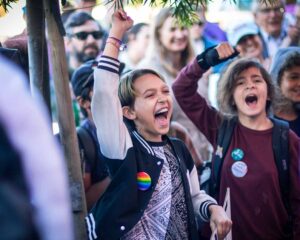 A student with a rainbow pin throws their hand up into the air and cheers. More students and families can be seen in the background cheering.