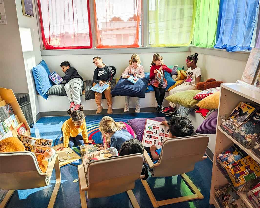 First-grade students sit and read in a library with colorful rainbow curtains in the windows.