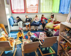 First-grade students sit and read in a library with colorful rainbow curtains in the windows.