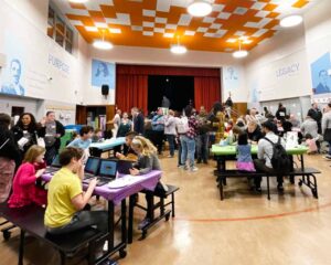 Groups of students and families in the school auditorium meet around tables playing math games.