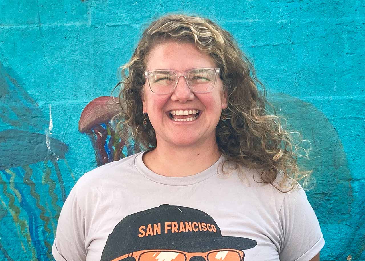 Ms. Summer smiles and stands in front of a blue mural.