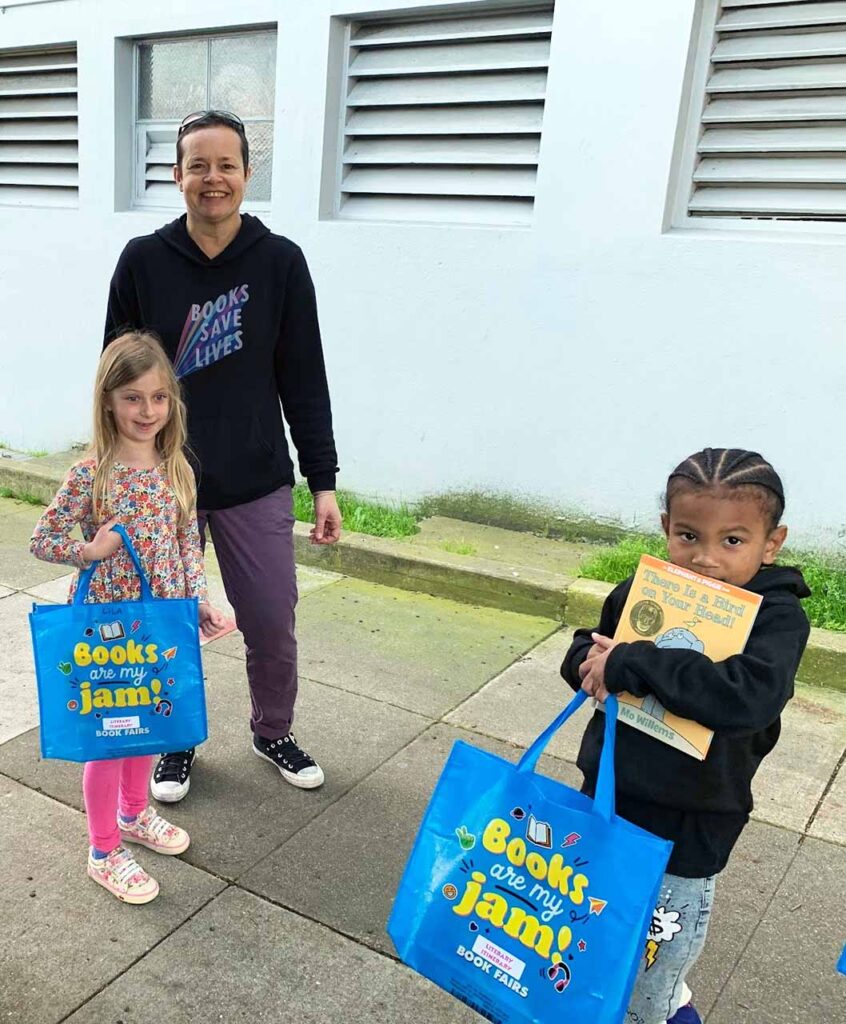 Ms. Sarah stands outside the school with two young students holding books and book bags.