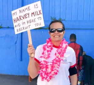Ms. Sarah, dressed up as Harvey Milk for Halloween. She wears a pink lei and holds a sign that reads “My name is Harvey Milk and I'm here to recruit you.”