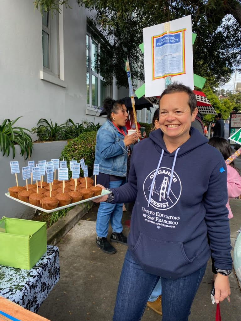Ms. Sarah outside the school at a picket, holding a tray of muffins.
