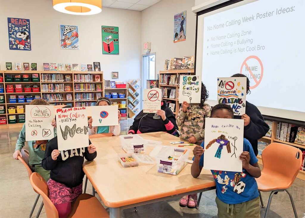 Students in the library hold of hand-drawn posters in front of their faces to promote "No Name Calling Week."