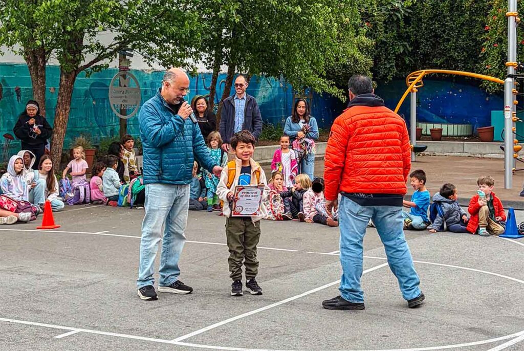 Mr. Sanchez and a young student at morning circle, with students and parents in the background.