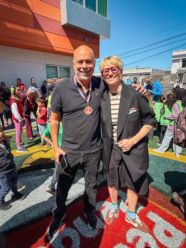 Principal Mark Sanchez stands with Megan Rapinoe at Harvey Milk Civil Rights Academy, surrounded by a crowd of students on the new soccer pitch.
